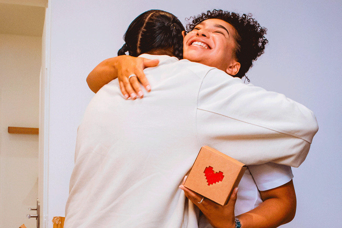 A couple embracing at home, one holding a Lovebox showing a pixel-heart message.